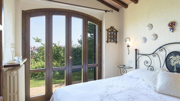 A bedroom with a large sliding glass door to a garden, a bed with white linens, a small bedside table with a cup, and a wall-mounted light fixture. - Tr-i381-vena0at - Casale al Doglio 6 1 (San Venanzo)
