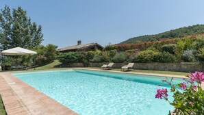 A rectangular swimming pool with clear blue water, surrounded by a stone deck. There are two lounge chairs and a covered patio area with a white canopy. The pool is set against a backdrop of lush greenery and a hillside. - Tr-i381-vena0at - Casale al Doglio 6 1 (San Venanzo)