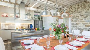 A modern kitchen with stainless steel appliances, a wooden dining table set for six, and a stone wall in the background. - Ar-a390-arez0bt - Torre Ginestre 12 (Arezzo)