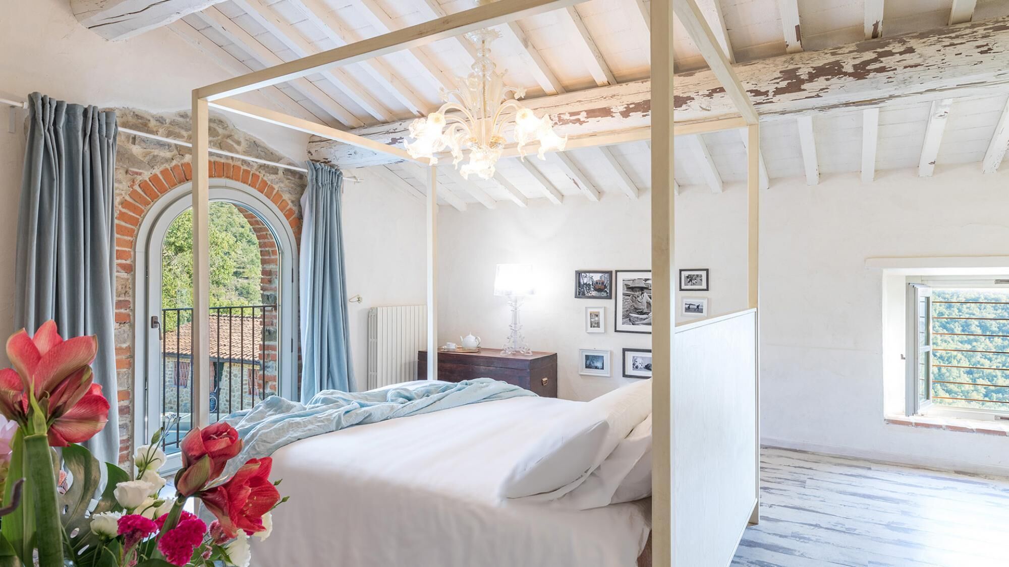 A bedroom with a four-poster bed, a chandelier, a dresser, and a window with a view of greenery.