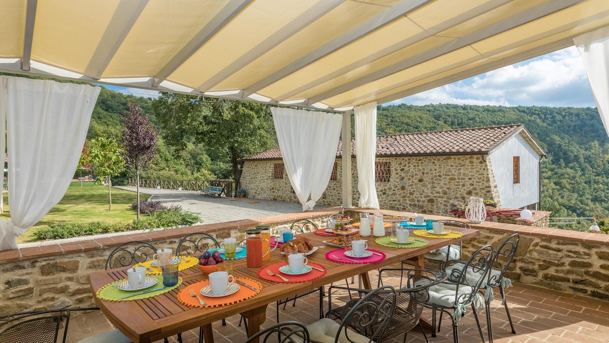 An outdoor dining area with a table set for a meal, surrounded by stone walls and a view of a stone house and forested hills.