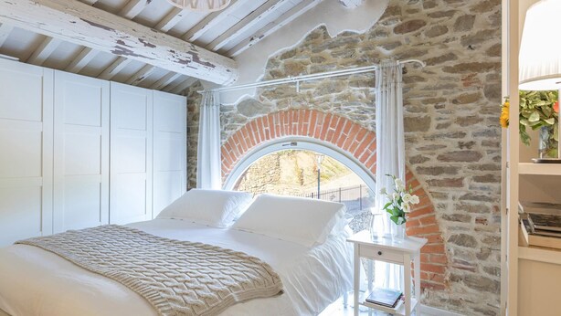 A bedroom with a stone wall, a white bed, and a small table with a book. - Ar-a390-arez0bt - Torre Ginestre 12 (Arezzo)