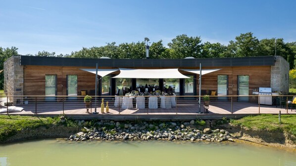 A modern outdoor dining area with a covered table and chairs, a stone wall, and a view of a river.