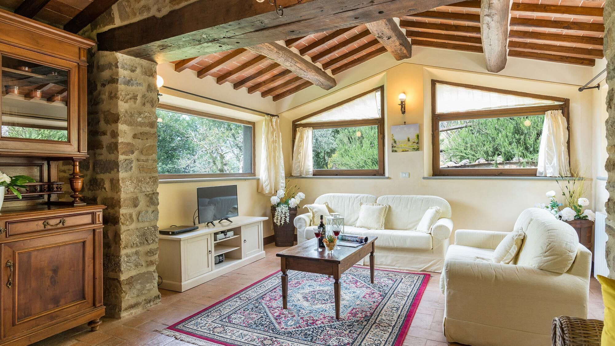 Una sala de estar acogedora con techo de madera, un sofá blanco, una mesa de centro y un televisor. Se observan ventanas con cortinas, una pared de piedra y un mueble de madera rústico.