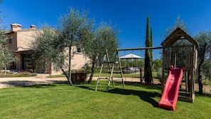 Un'area verde esterna con altalene in legno, uno scivolo rosso e una casetta gioco in legno.