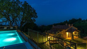 A swimming pool with a metal railing, a stone house with a tiled roof, and a view of the surrounding landscape at dusk.