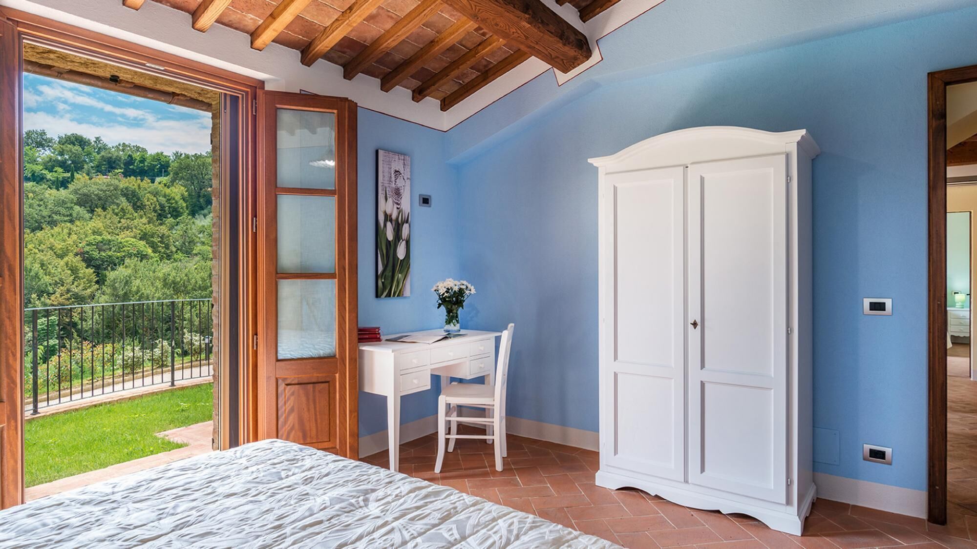 A bedroom with a bed, a desk, a chair, a wardrobe, and a balcony with a view of greenery.