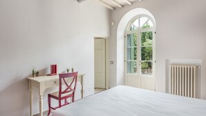 A bedroom with a white bed, a desk with a red chair, a radiator, and a window with a view of greenery.
