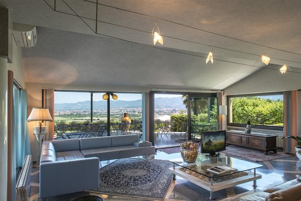 A spacious living room with a large sofa, a coffee table, and a television. There is a view of the city and mountains through large windows.