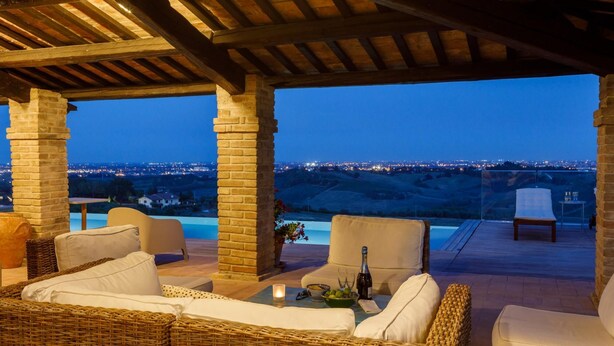A covered outdoor seating area with wicker furniture, a table with a bottle and glasses, and a view of a cityscape at dusk. - Villino Bruna 6 in Bertinoro (Bertinoro)