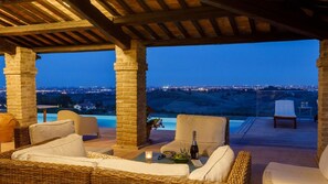 A covered outdoor seating area with wicker furniture, a table with a bottle and glasses, and a view of a cityscape at dusk.