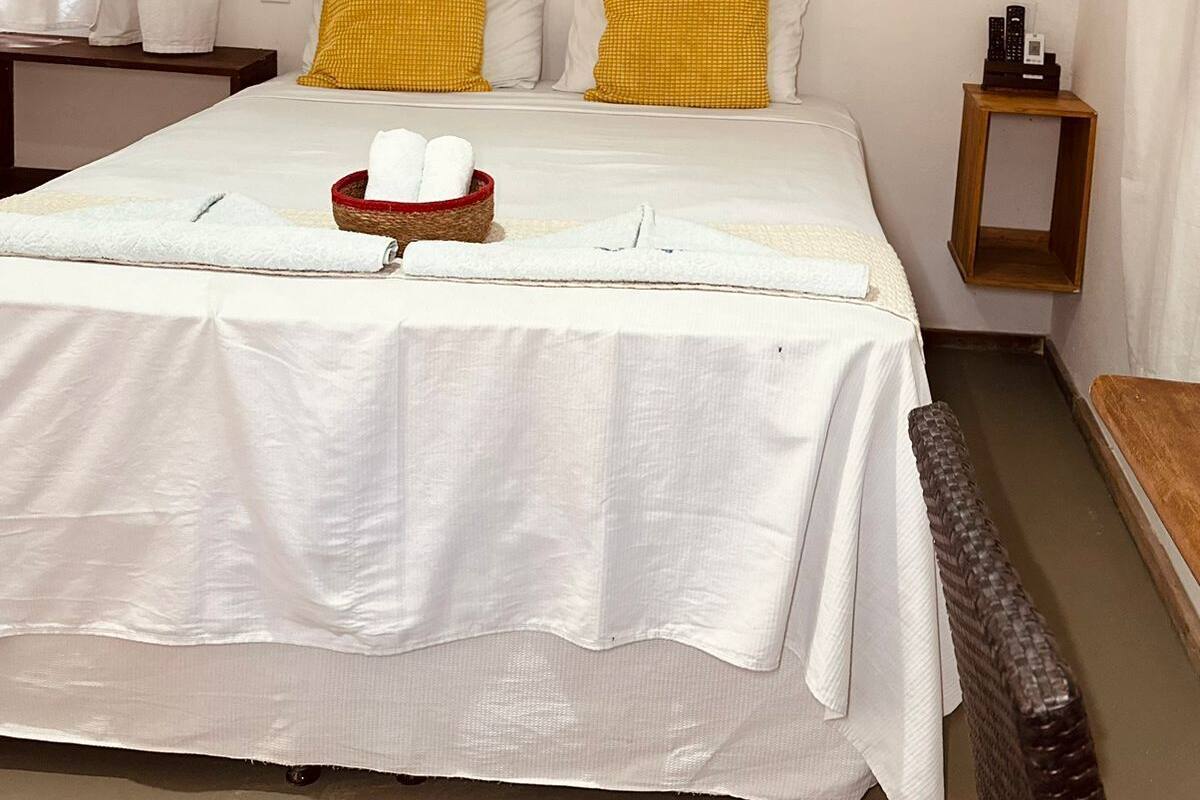 Ein ordentlich gemachtes Bett mit weißen Bettwäsche, zwei gelben Kissen und einem kleinen Korb mit weißen Handtüchern.