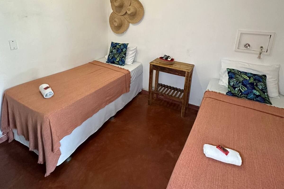Ein Zimmer mit zwei Einzelbetten, jeweils mit einer braunen Bettdecke und einem weißen Kopfkissen. Es gibt einen kleinen Holztisch mit einem Korb darauf und eine an der Wand befestigte Hutfassung.