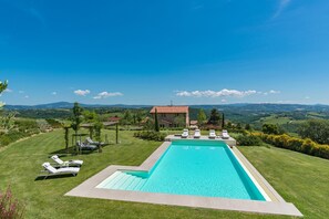 A rectangular swimming pool with a clear blue water, surrounded by a well-maintained lawn and white lounge chairs. In the background, there is a stone building with a red roof, and a view of rolling hills and distant mountains under a clear blue sky.