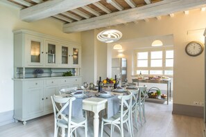 A dining area with a white table set for four, surrounded by white chairs. There is a kitchen area with white cabinets and a refrigerator.