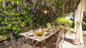 An outdoor dining area with a long table set for a meal, surrounded by lush greenery and purple flowers. - Le Querce Dei Medici 12 2 (Scarperia e San Piero)