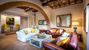 A spacious living room with a white sofa, a brown leather sofa, and yellow armchairs. There is a coffee table with magazines and a vase of flowers. The room has exposed wooden beams and a large mirror on the wall. - Le Querce Dei Medici 12 2 (Scarperia e San Piero)