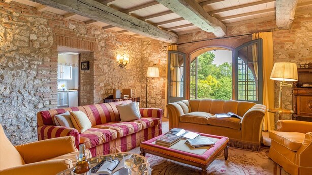 A cozy room with stone walls, a wooden ceiling, and a large window offering a view of greenery. There are two sofas and a coffee table with books and glasses.