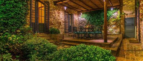 A stone building with ivy-covered walls, a wooden pergola, and a table with chairs in the center.