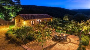 A stone house with a tiled roof, surrounded by a garden with a table and chairs, and a view of a forested hill. - Muffato 6 in Sarteano (Sarteano)