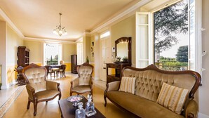 Ein geräumiges Wohnzimmer mit einer großen Couch, zwei Sesseln, einem Couchtisch und einem Chandelier. Ein Essbereich mit Tisch und Stühlen lädt zum Verweilen ein, und durch ein großes Fenster bietet sich ein Blick auf den Garten.