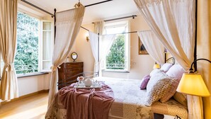 Ein Schlafzimmer mit Bett, Nachttisch, Lampe und Fenstern mit Vorhängen.