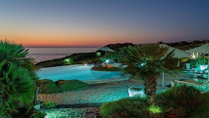 A resort pool area with a view of the ocean at sunset, surrounded by lush greenery and outdoor seating. - Villa Ambra 10 in Sorso (Castelsardo)