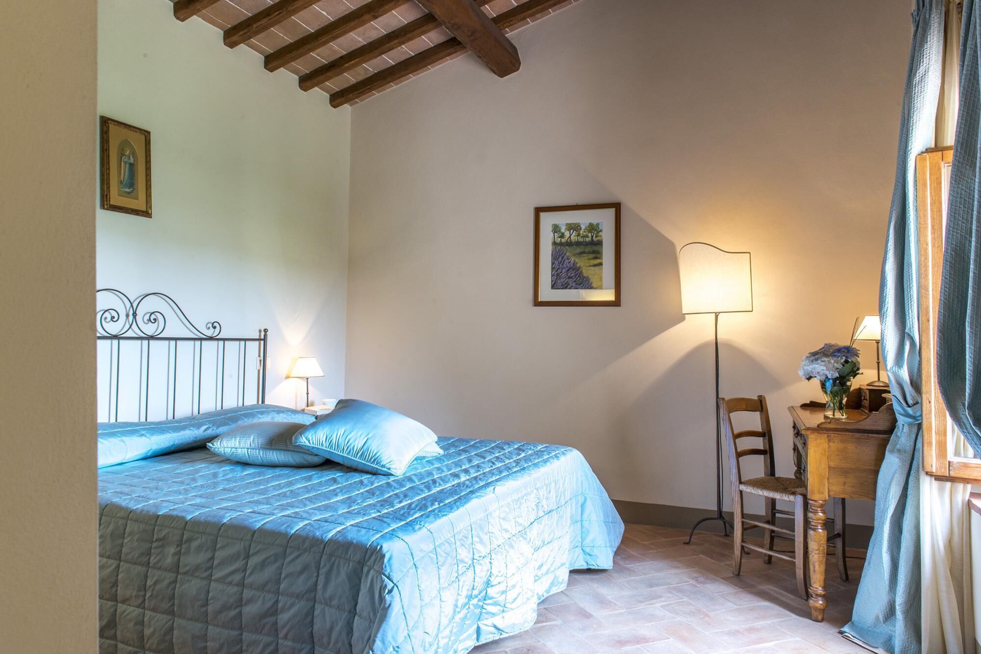 Un dormitorio con cama, escritorio, silla, lámpara y un cuadro en la pared.