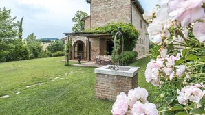 Un bâtiment rustique en pierre, doté d’un patio couvert et d’un jardin agrémenté de fleurs roses.