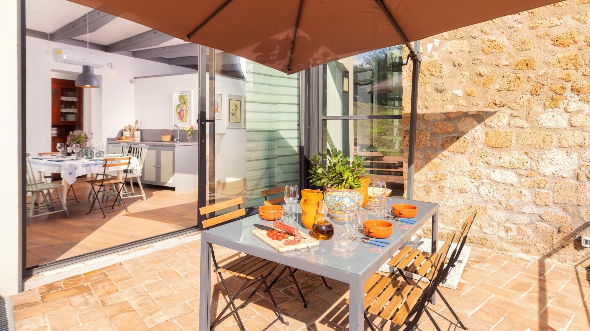 An outdoor dining area with a table set for two, a potted plant, and a stone wall in the background.