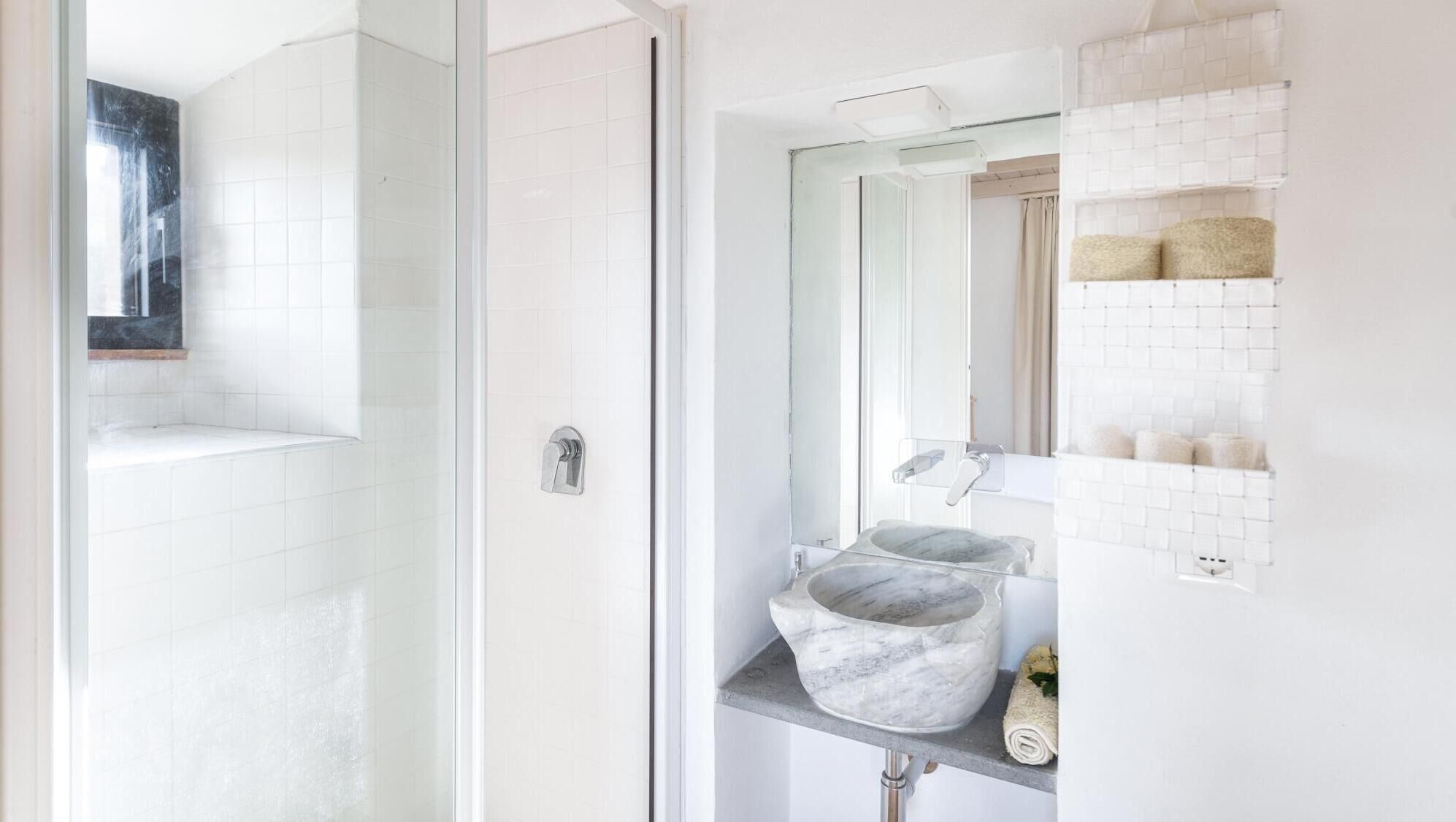 A bathroom with a glass shower enclosure, a marble sink, and a tiled wall with a shelf holding toiletries.