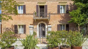 A brick building with a central entrance, multiple windows with shutters, and a metal gate. There are potted plants in front of the building.