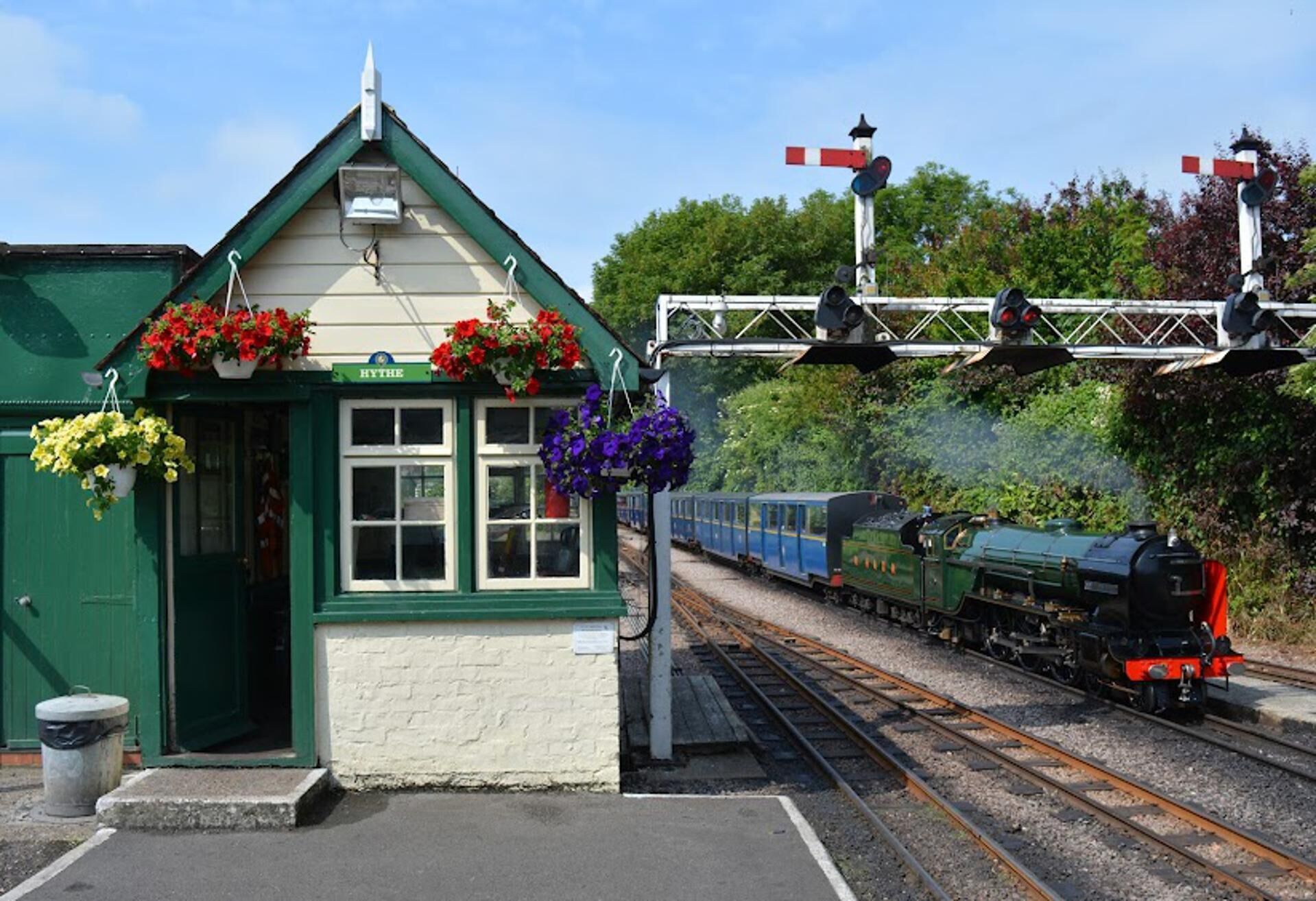 A small station building with a green roof and flower boxes, a railway track with a steam locomotive, and a level crossing with barriers.