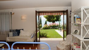 A cozy living room with a sofa, a chair, and a bookshelf. A large window offers a view of a garden and a body of water.
