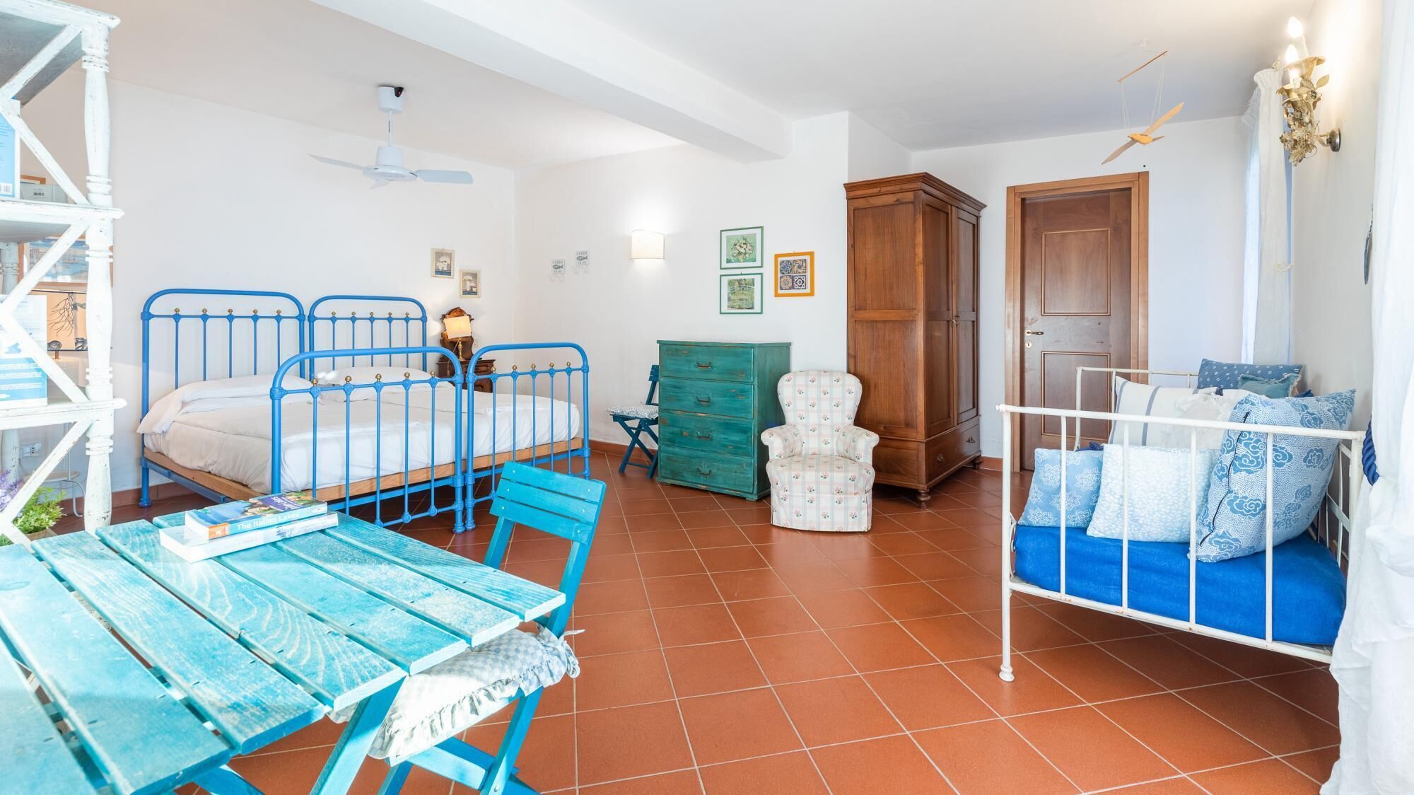 A room with two beds, a wooden wardrobe, a green chest of drawers, a white chair, and a blue table.
