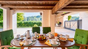An outdoor dining area with a table set for breakfast, surrounded by green chairs, and a view of a garden and hills in the background. - Cascina La Valle 10 2 (Ponzano Monferrato)