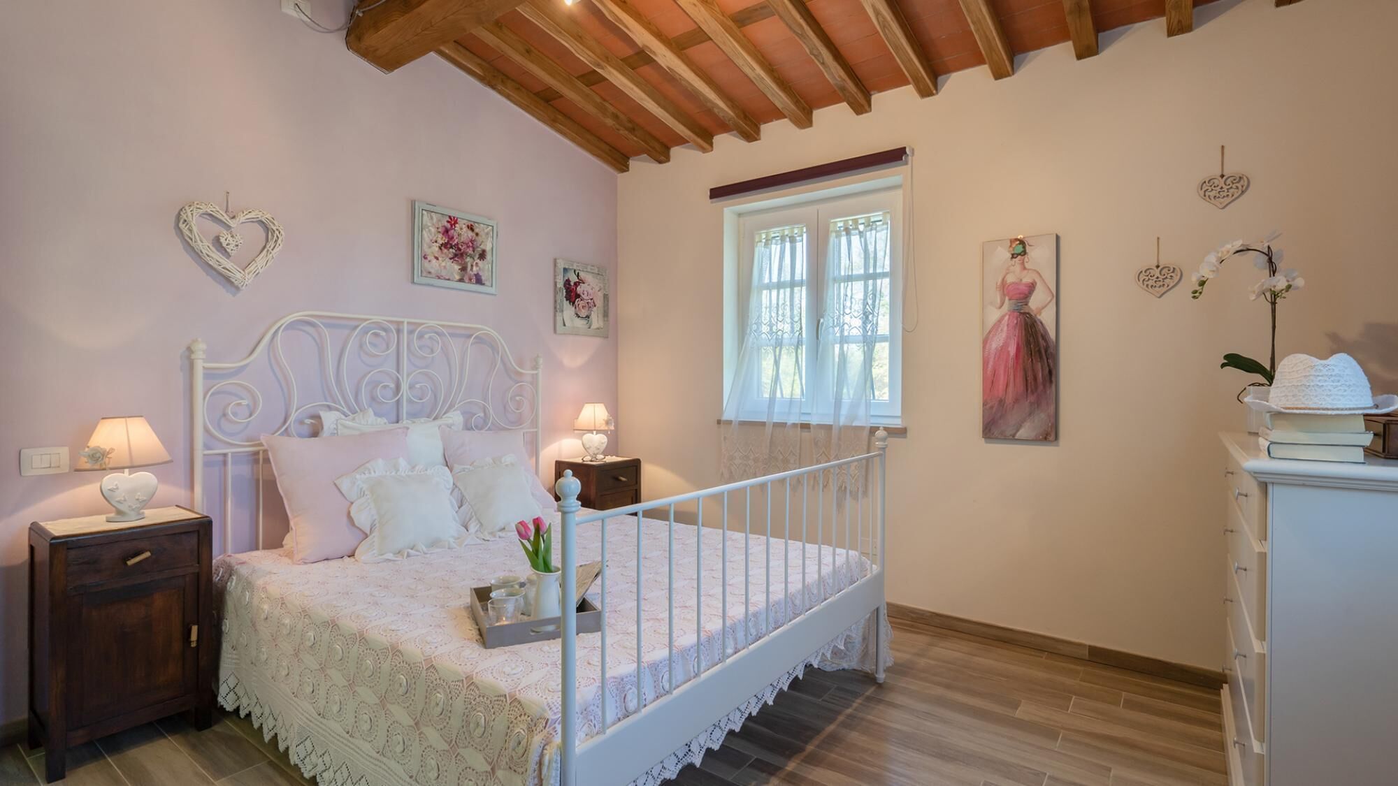 A bedroom with a white bed, wooden headboard, two bedside tables with lamps, a window with sheer curtains, and a dresser with a hat on top.