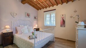 A bedroom with a white bed, wooden headboard, two bedside tables with lamps, a window with sheer curtains, and a dresser with a hat on top. - Ai Due Cuori 4 2 in Capannori (Capannori)