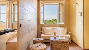 A room with a wooden floor, a sofa, a small table, and a window with a view of greenery. - Casa Del Vento 10 in Bibbona (Bibbona)