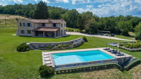 A stone house with a swimming pool, surrounded by well-maintained lawns and a forested backdrop.