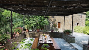 Un patio al aire libre con mesa de madera lista para comer, rodeado de muros de piedra y plantas en macetas.