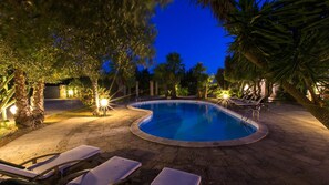 Ein Pool mit gewölbter Kante, umgeben von Liegestühlen und Palmen, im Abendlicht.