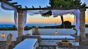 Area esterna coperta con sedute, cuscini bianchi e vasi in terracotta, affacciata su una vista costiera.