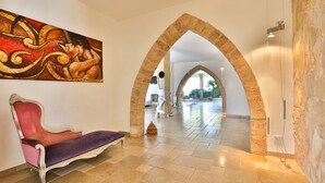 Una hall con una poltrona in velluto rosso, un grande quadro astratto e un arco che conduce a un'ampia zona con pavimento in pietra e un lampadario.