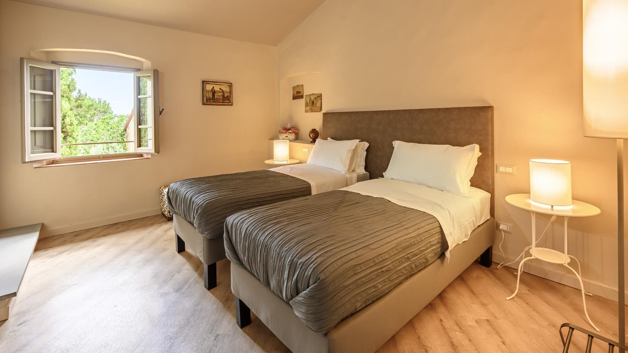 A bedroom with two beds, a window, a bedside table with a lamp, and a small table with a vase.