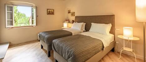 A bedroom with two beds, a window, a bedside table with a lamp, and a small table with a vase.