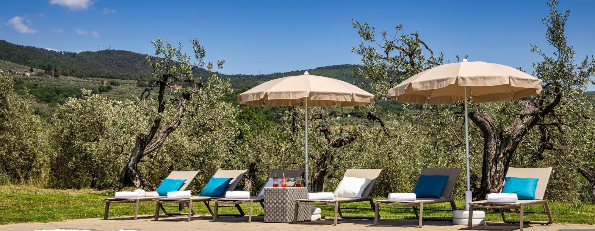 A row of sun loungers with beige cushions and blue pillows, shaded by beige umbrellas, set against a backdrop of olive trees and a hilly landscape.