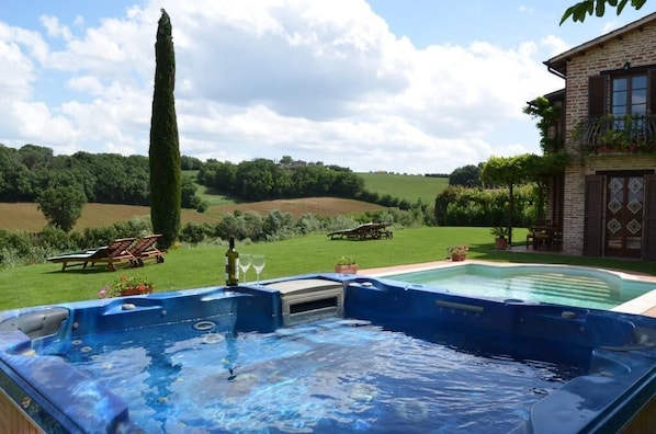 A hot tub in a backyard with a pool, surrounded by a house and a grassy field.