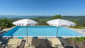 A rectangular swimming pool with clear blue water, surrounded by lounge chairs and white umbrellas, set against a backdrop of a mountainous landscape and a clear sky. - Antiche Pietre 10 in Tuoro sul Trasimeno (Tuoro sul Trasimeno)