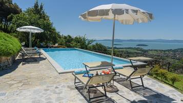 Piscina retangular com espreguiçadeiras e guarda-sóis, cercada por pavimento de pedra e vegetação, com vista para uma paisagem deslumbrante.